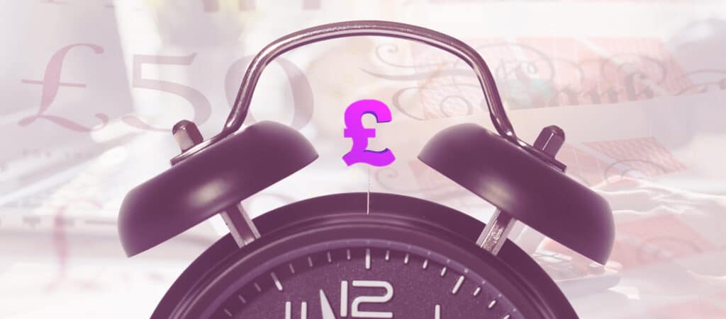 A traditional wind-up or key-wound, mechanical spring-powered alarm clock with two bells, between the bell is a pound sign. In the background there are fifty pound notes and and laptop - so show that time is money.