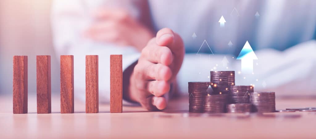 A hand blocking wooden blocks from falling towards stacked coins, illustrating credit risk reduction strategies for a recruitment agency.